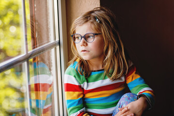 Adorable little preschool girl with eyeglasses sitting by the window. Thoughtful child looking out. Lonely kid.