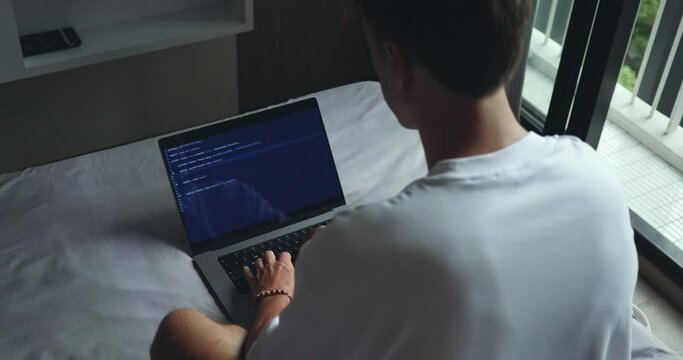 Digital Workspace. Man Typing Programmer Code on Laptop, Seated on Bed, Rear View. IT Freelancer writing code on his laptop from the comfort of his bed