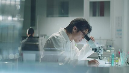 View through glass wall of Asian scientist in lab coat sitting at desk, looking through microscope and writing down notes during laboratory research - Powered by Adobe