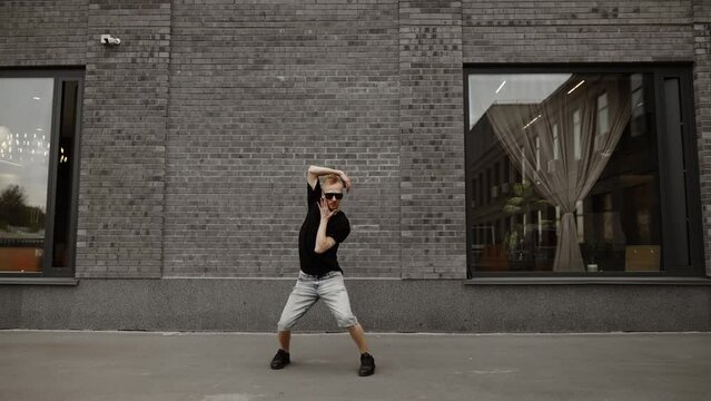 A dancer in gray shorts and a black T-shirt with glasses dances against the background of a gray brick wall. Slow motion, dynamic camera hitting.
