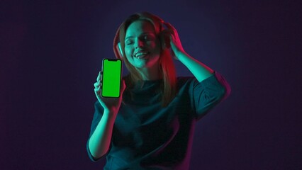 A woman in red wireless headphones shows a green smartphone screen. A woman listens to music with her eyes closed, using an app, in a studio on a blue background in green and pink neon light.