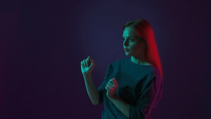 Fototapeta premium A young woman works on an invisible touch screen. A woman in the studio on a blue background in pink and green neon light. Futuristic technology.