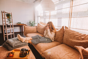 Happy woman relaxing on sofa at home on bright winter morning - 649612825