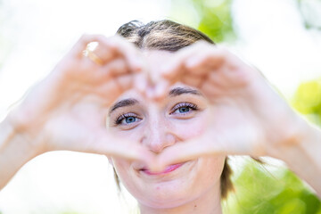 In this captivating close-up, a young woman holds her hands up in a heart shape, framing her face...