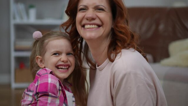 Positive woman talking as girl covering lips with finger and lady biting kid laughing out loud. Side view portrait of joyful carefree Caucasian mother and daughter having fun enjoying weekend at home
