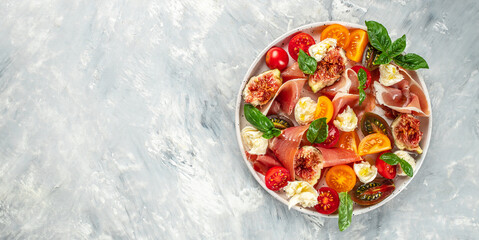 Healthy and delicious salad with mozzarella, figs, prosciutto, fresh basil leaves and tomato on a light background. Long banner format. top view