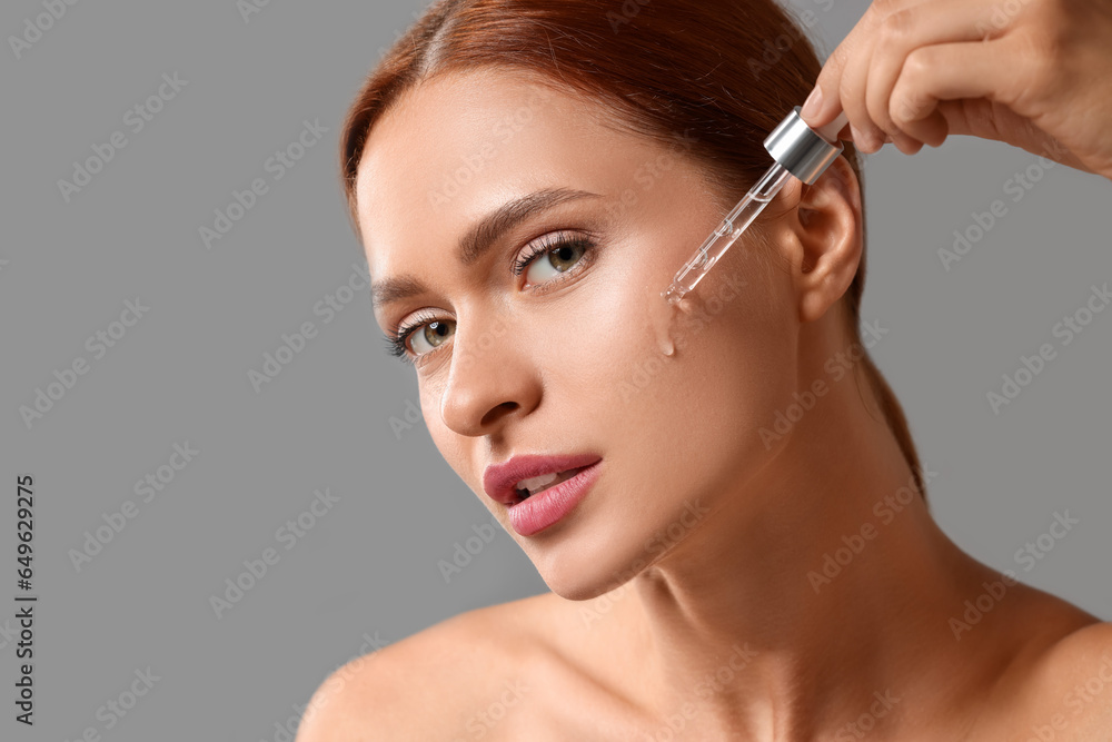 Wall mural beautiful young woman applying cosmetic serum onto her face on grey background, space for text