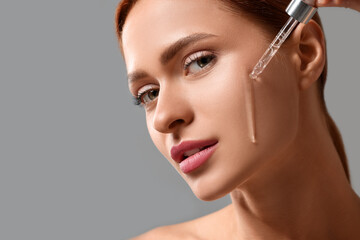 Beautiful young woman applying cosmetic serum onto her face on grey background, closeup. Space for...
