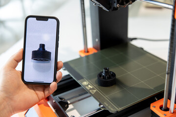 Man holding smartphone with detail on screen, working on 3D printer in creative lab.