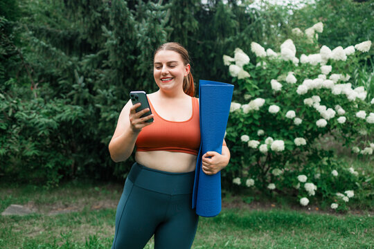 Young 20s fat woman hold yoga mat and using smartphone after workout on street. Portrait of smiling female in sportswear walk using smartphone. High quality photo