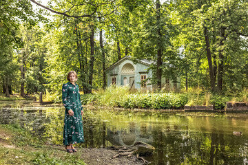 A blonde woman in a summer green floral dress stands on the bank of the river in an old manor....