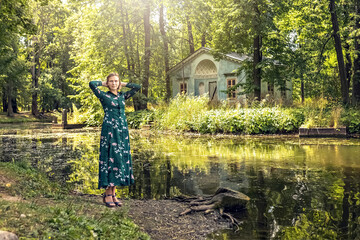 A blonde woman in a summer green floral dress stands on the bank of the river in an old manor....