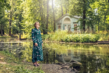 A blonde woman in a summer green floral dress stands on the bank of the river in an old manor....