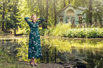 A blonde woman in a summer green floral dress stands on the bank of the river in an old manor....