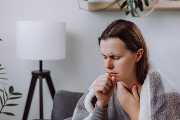 Upset sick young woman touching painful swollen neck glands, suffering from throat infection, unhealthy sad caucasian female covered plaid sitting on couch at home feeling pain with swallowing - obrazy, fototapety, plakaty