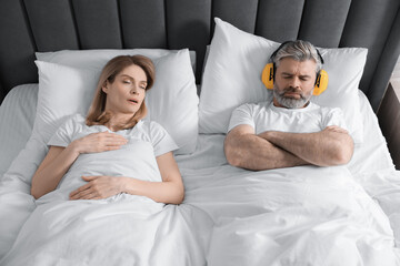 Irritated man with headphones lying near his snoring wife in bed at home