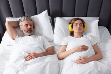 Smiling woman with headphones lying near her snoring husband in bed at home