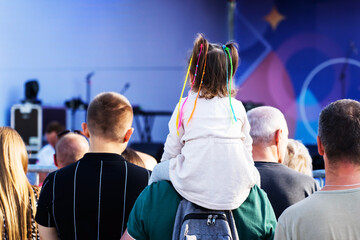 A little girl with colorful pigtails sits on her father's shoulders during a mass outdoor concert....