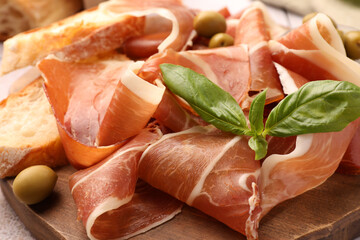 Slices of tasty cured ham, olives, bread and basil on wooden board, closeup