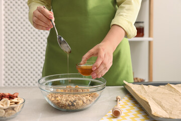 Making granola. Woman adding honey into bowl with mixture of oat flakes and other ingredients at light marble table in kitchen, closeup