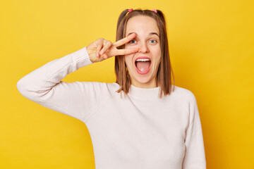 Excited amazed teenage girl in jumper with ponytails isolated over yellow background celebrating her victory success showing v sign in front her eye yelling with happiness.