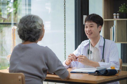 Friendly Smiling Young Asian Doctor Talking And Explaining Test Results To An Elderly Patient In A Hospital.
