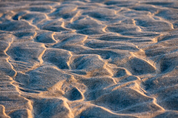 Fototapeta na wymiar Sand structures with irregular ripples at low tide on the beach of Juist island, Germany in National Park “Wattenmeer“. Natural background pattern formed by water current and wind, selective focus.