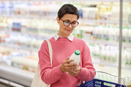 Waist up portrait of mature woman holding bottle of milk in supermarket and looking at expiration date, copy space