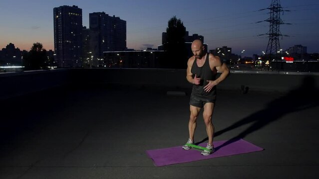 A man with an athletic build is doing sports on a rooftop in a night city. The athlete places his feet shoulder-width apart and raises the leg bent at the knee. High quality 4k footage