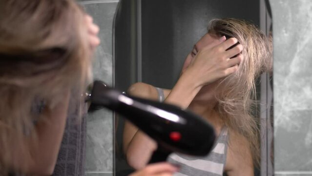 Hair care. Blonde Woman Drying Styling Hair Blow Dryer Creating Hairstyle Standing in Bathroom At Home. Woman using hair dryer after morning shower. Household routine in morning when going to work.