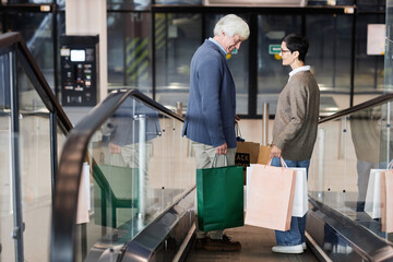 Fototapeta premium Side view of senior couple holding shopping bags standing on escalator in shopping mall, copy space