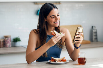 Beautiful sporty woman eating healthy sanwich while using smartphone in the kitchen at home