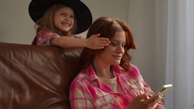 Concentrated woman scrolling smartphone touchscreen as cheerful blurred girl in Halloween witch costume covering eyes with hands. Happy Caucasian mother and daughter laughing having fun indoors