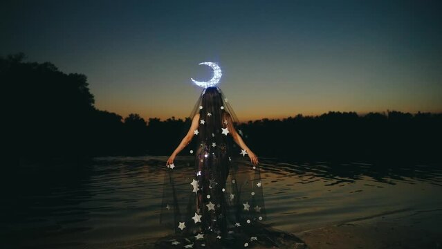 Fantasy woman queen of night lady moon, crown glows on head, black dress cape shining stars back rear view. fairy girl princess creative costume. Summer dark nature water lake river sky sunset. 4k