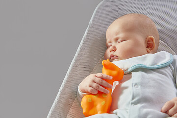 The baby sleeps with an orange toy rubber giraffe in a chaise longue on a gray background with...