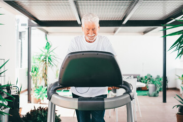 People sport concept. Happy senior man doing exercises to stay fit. Elderly bearded man in sportswear running on treadmill in home terrace - Powered by Adobe