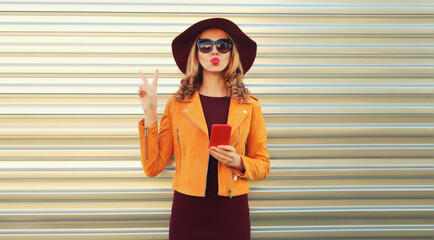 Portrait of stylish young woman posing blowing her lips sends sweet air kiss holds phone wearing sunglasses, round hat and jacket on gray background