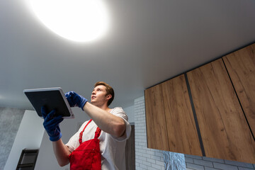 Worker installing lamp on stretch ceiling indoors.