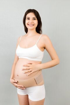 Portrait of supporting bandage on pregnant woman in underwear at gray background with copy space. Orthopedic abdominal support belt concept