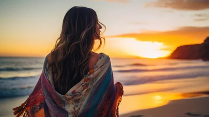 Foto op Plexiglas Strand zonsondergang Young beautiful woman looking at sunset on the beach with a shawl on her shoulders on a fresh evening