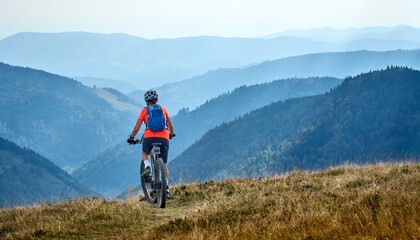 nice senior woman on her electric mountain bike cycling on Feldberg summit with stunning view over the Black Forest mountains and valleys, Baden-Wuerttemberg Germany - obrazy, fototapety, plakaty