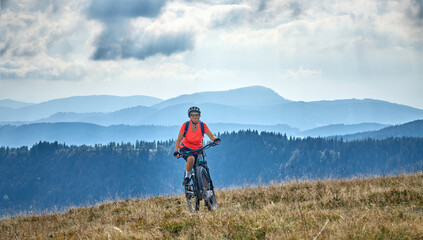 Fototapeta premium nice senior woman on her electric mountain bike cycling on Feldberg summit with stunning view over the Black Forest mountains and valleys, Baden-Wuerttemberg Germany