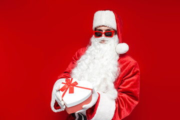 santa claus in glasses holds gift box in the shape of heart on colored background, man in hat and santa costume
