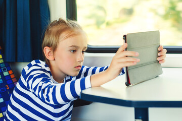 Kid travels on a train. Journey. Gadget addiction. Cute child playing video games online on tablet during trip.