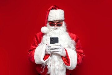 santa claus in hat and festive glasses uses smartphone on red background, man in santa costume types online