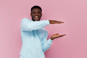 african american man in blue sweater holds empty space on pink isolated background, the guy shows...