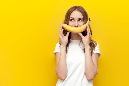 young girl shows smile and holds banana on yellow isolated background, woman with fruit looks at copy space