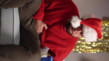 a man on the sofa in a Santa Claus hat, holding a bucket in his hands and holding his stomach, he is vomiting. concept of food poisoning during Christmas or New Year. close-up portrait.