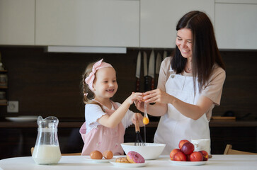 Mother and daughter preparing a sweet cake using flour, milk, sitting on chairs at a table in a...