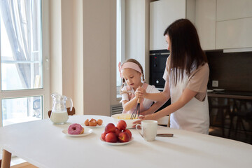 Mother and daughter preparing a sweet cake using flour, milk, sitting on chairs at a table in a...